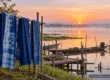 AI-generated conceptual scenery inspired by Sakon Nakhon: indigo-dyed cloth drying on bamboo poles beside a misty lake and fishing boats at sunrise (not a photograph of a real place)