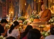 Devotees kneeling with offerings before a seated monk image in a candle-lit, flower-filled Thai temple hall. AI-generated image.