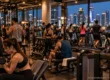 Crowded gym packed with exercise machines and people working out at night, the Bangkok skyline through the windows. AI-generated image.
