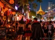 Costumed crowd at a Halloween street party in Bangkok at night, with pumpkin lanterns, a golden temple and the BTS Skytrain in the background. AI-generated image.