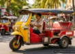 Red and yellow tuk-tuk with its driver on a sunny Phuket street lined with palm trees and shops, with more tuk-tuks in the background. AI-generated image.
