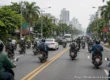 Busy multi-lane road in Thailand filled with helmeted motorbike riders and cars, seen from behind the traffic with palm trees and city towers ahead. AI-generated image.