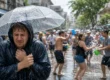 Drenched tourist in a black raincoat sheltering under a clear umbrella while a lively Songkran water fight carries on in the street behind him. AI-generated image.