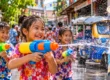 Laughing Thai children with water guns splashing during Songkran, with a tuk-tuk on the street behind them. AI-generated image.