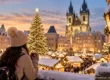 Traveller with a hot drink overlooking a snowy European Christmas market with a lit tree and Gothic church at dusk. AI-generated image.